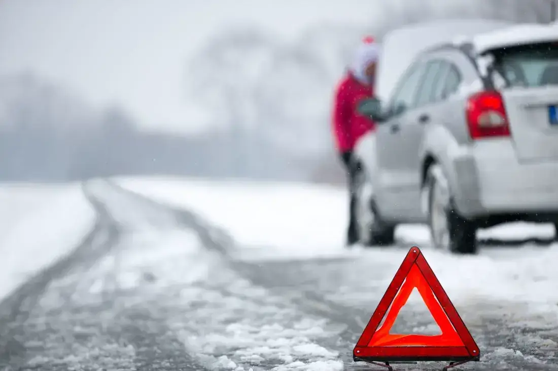 Ein Warndreieck steht vor einem Auto mit geöffneter Motorhaube und einer Person nach einem Unfall mit Sommerreifen im Winter.