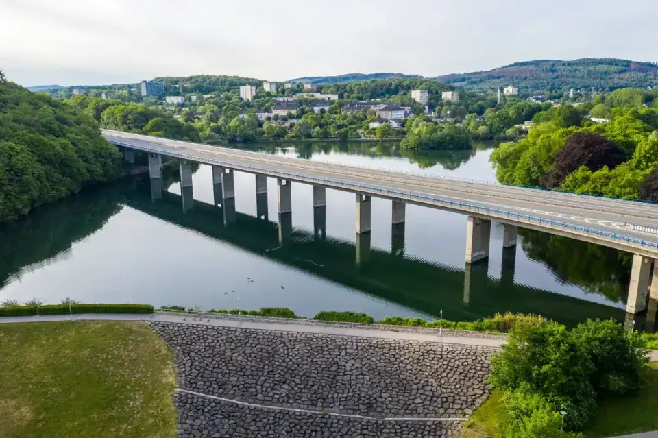 Die Brücke über dem Seilersee in Iserlohn mit Wald, Gebäuden und Kfz-Werkstätten im Hintergrund. 